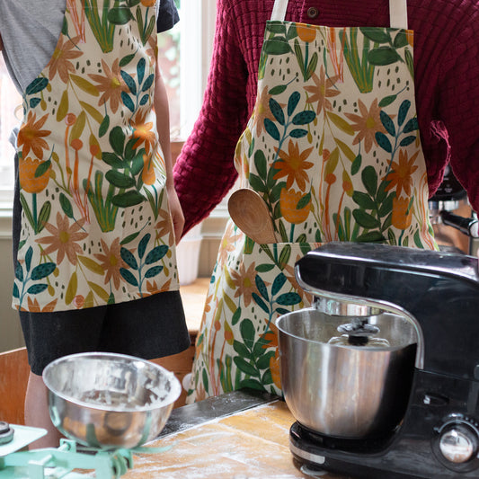 Child and adult matching aprons in a floral bud design. Adult apron has a pocket on the front and child's apron has an adjustable neck strap.Made in Britain.