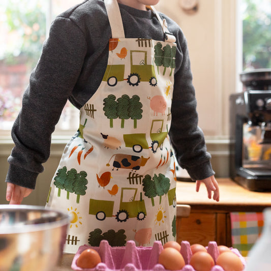 A child wearing a beige apron with green, yellow, and orange farm animals and tractors. 100% cotton. Adjustable neck strap and made in Britain.