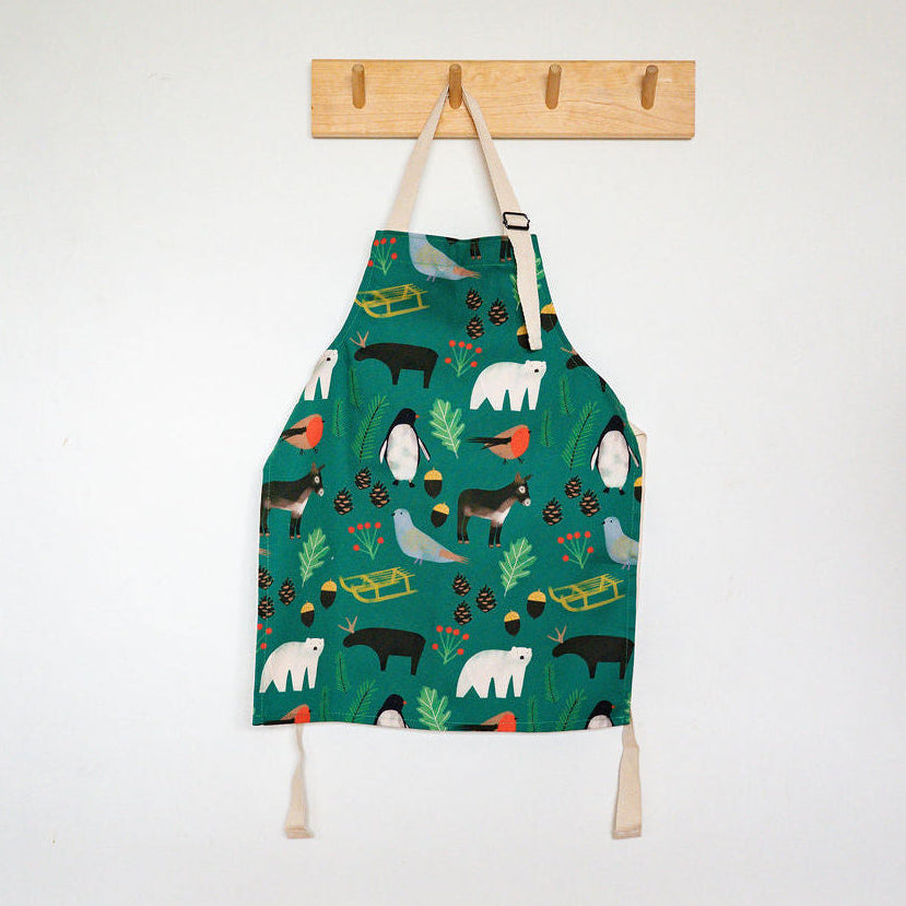 A Christmas children's apron with a colourful animal and nature print hangs from a wooden peg on a white wall above a wooden floor.