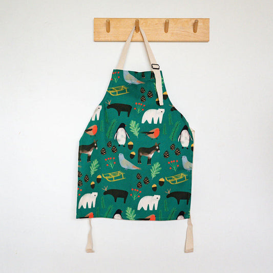 A Christmas children's apron with a colourful animal and nature print hangs from a wooden peg on a white wall above a wooden floor.