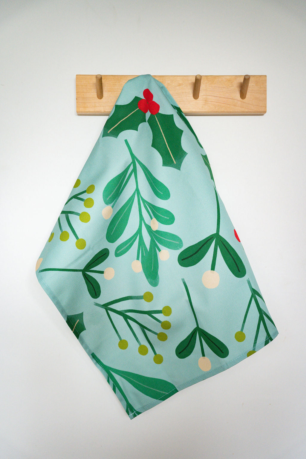 A light blue kitchen towel with a green mistletoe and holly pattern, hanging from a wooden peg on a white wall.