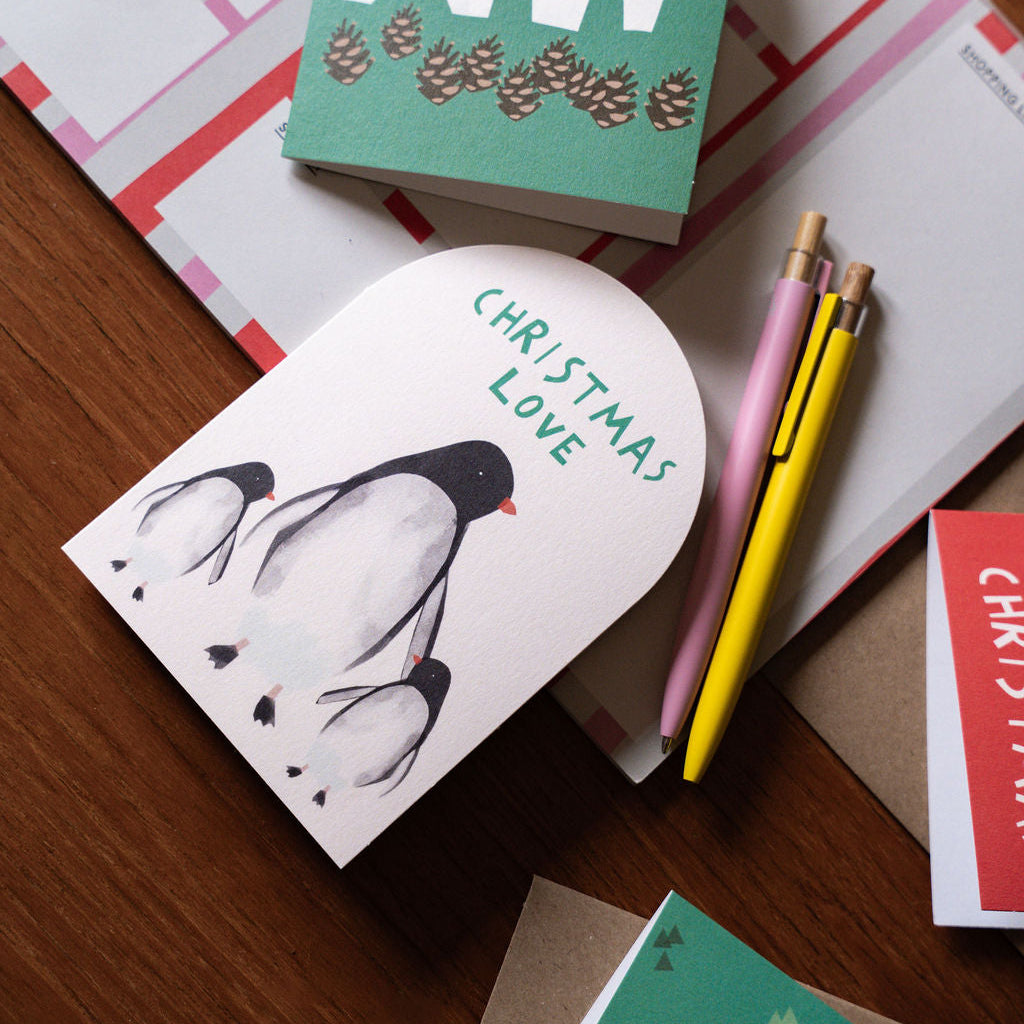 A styled flat-lay of Christmas greeting cards on a wooden table, featuring a green card with a white polar bear and pinecones reading 'Happy Christmas' and a white card with a black-and-white penguin reading 'Christmas Love'. Pink and yellow pens and envelopes are placed around them.