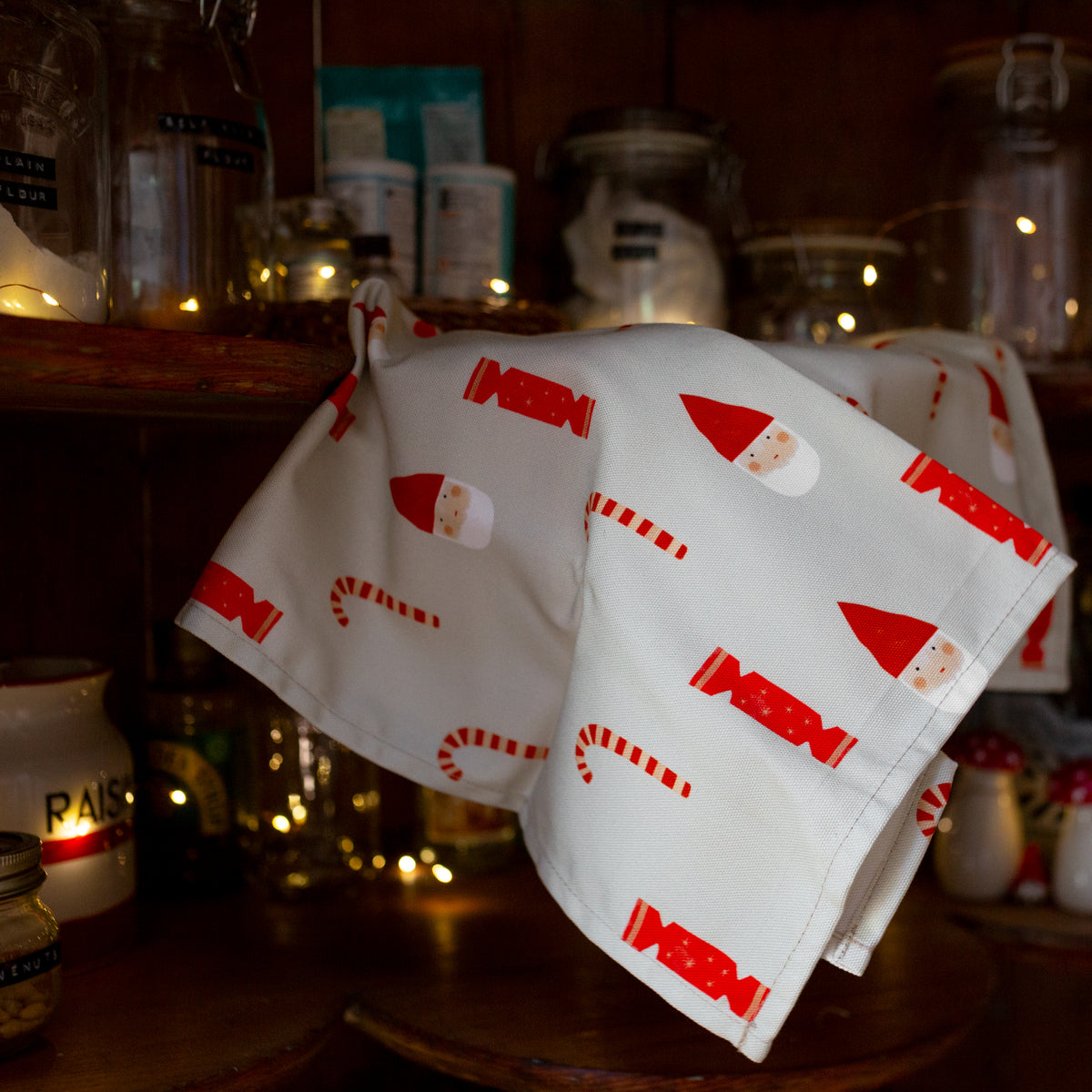 A white cotton tea towel with a limited edition Santa Claus design, hanging on a wooden rack in a home setting, decorated with Christmas lights and bottles in the background.