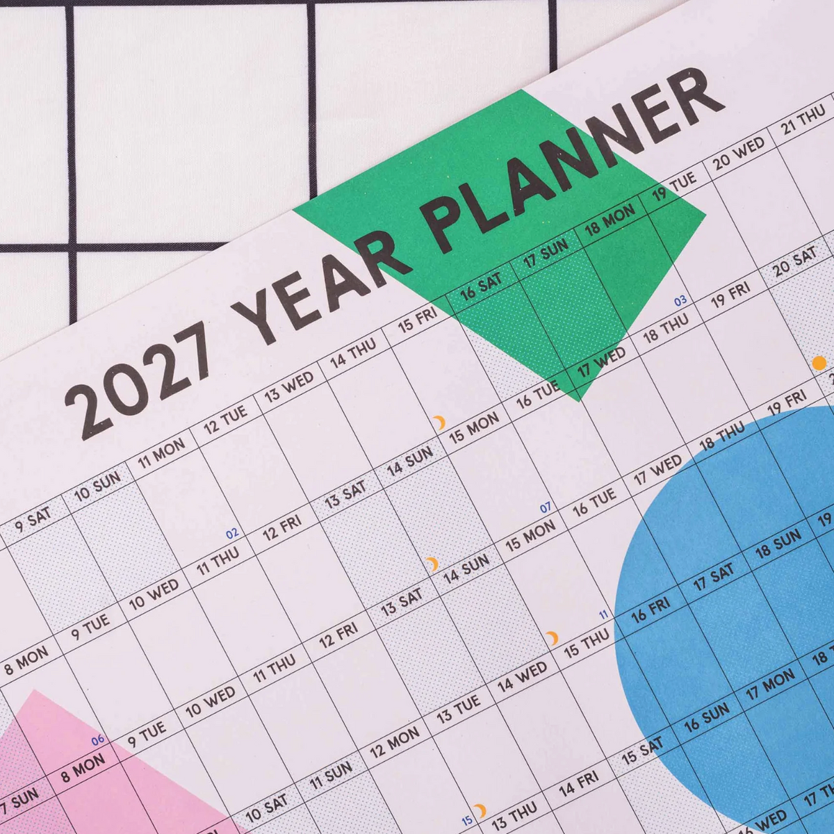 A close-up of a 2027 year planner page showing a monthly calendar grid with colored geometric accents in green, blue, and pink on a white background.