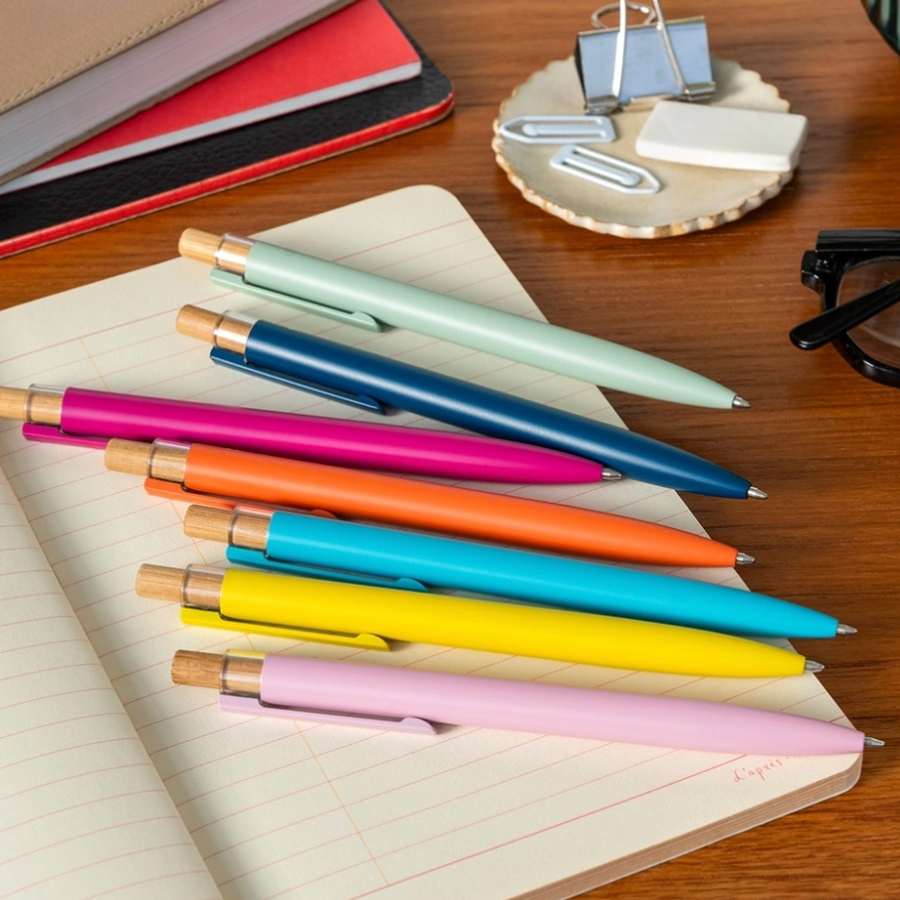 A set of eight colorful ballpoint pens with gold-toned metal accents arranged on an open lined notebook on a wooden desk, alongside other office items.
