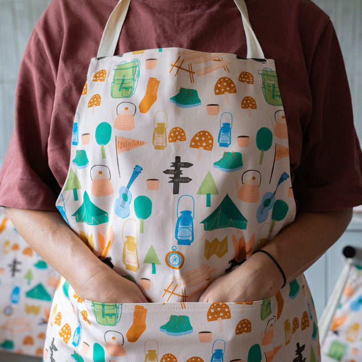 A person wearing a cream apron with a colorful camping-themed pattern of tents, campfires, and gear, paired with a maroon t-shirt in a kitchen setting.