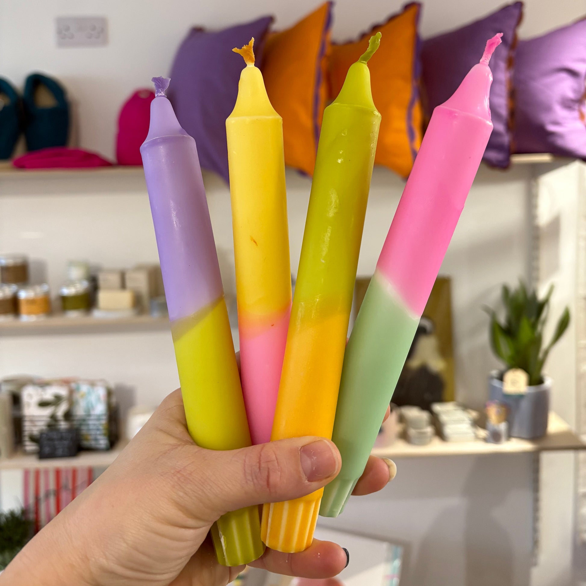 Hand holding four colorful candles in a store setting with shelves and pillows in the background.