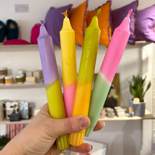 Hand holding four colorful candles in a store setting with shelves and pillows in the background.