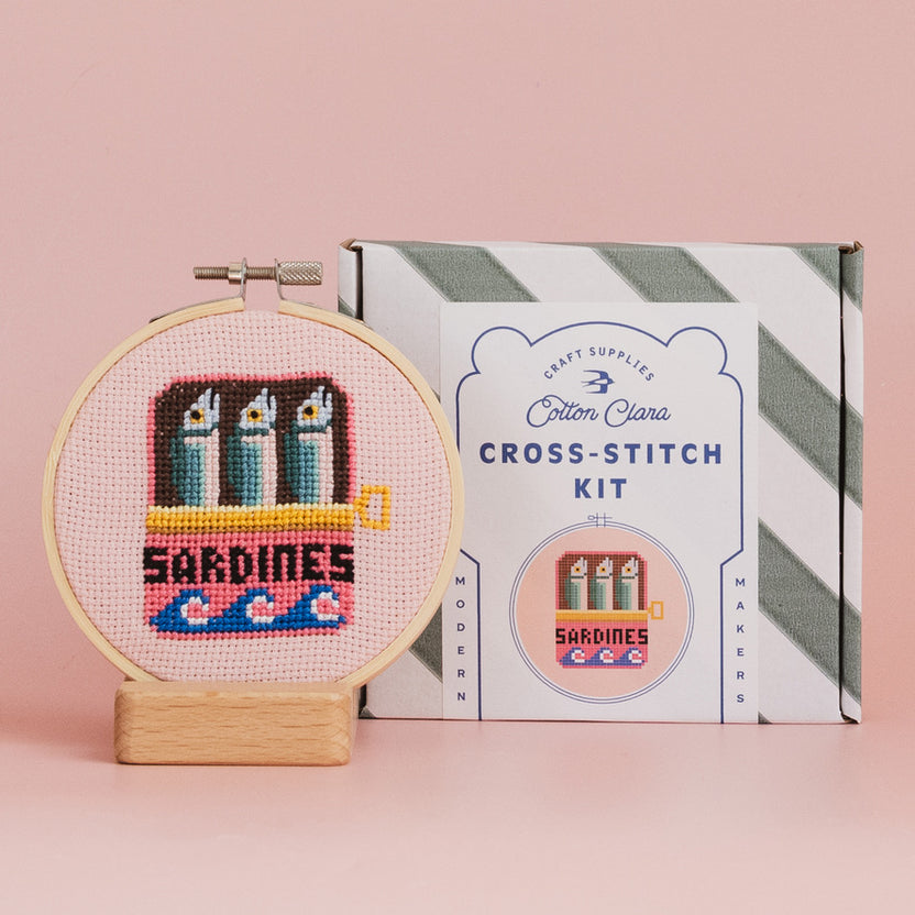 Cross-stitch kit with completed design of sardines in a can, next to its packaging on a pink background.