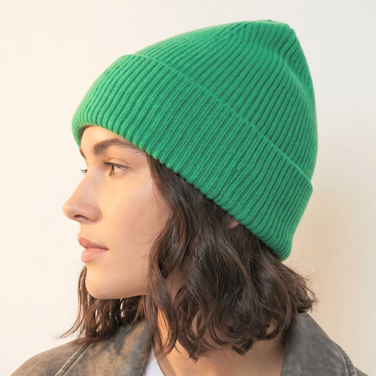 A woman in profile wearing a bright green ribbed knit beanie hat and a gray jacket over a white shirt, photographed against a plain light background.