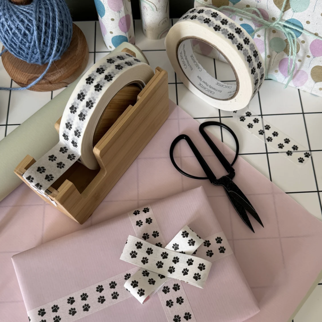 A styled flat-lay of washi tape with a black paw print pattern on a white background. The tape is shown in multiple rolls, some resting on a wooden display stand, with one roll wrapped around a gift box tied with a matching ribbon.
