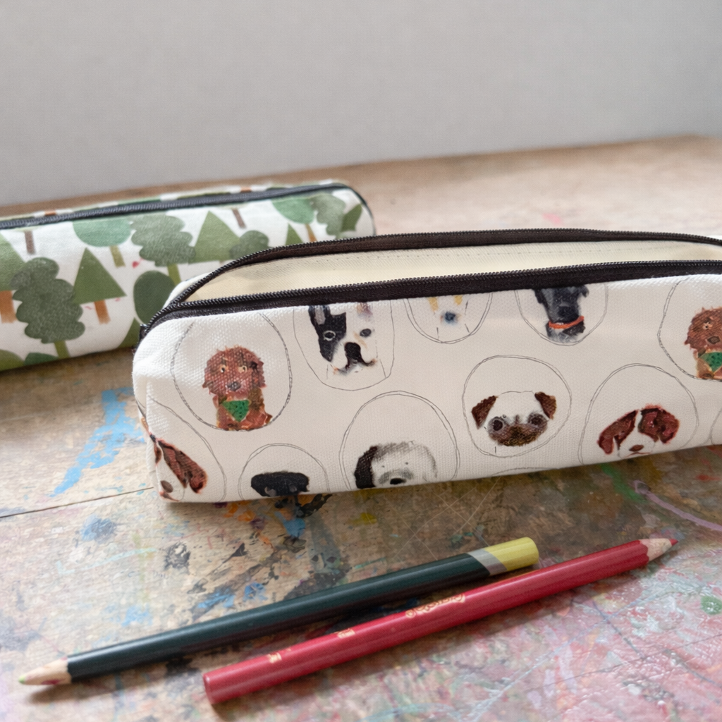A rectangular pencil case with a cream background and a pattern of various dog faces inside circles, shown alongside two colored pencils on a paint-splattered tabletop.