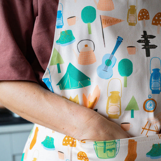 A close-up of a person wearing a pink apron with a colourful camping themed print, including tents, trees, lanterns, and a guitar, with their arm tucked into the front pocket.