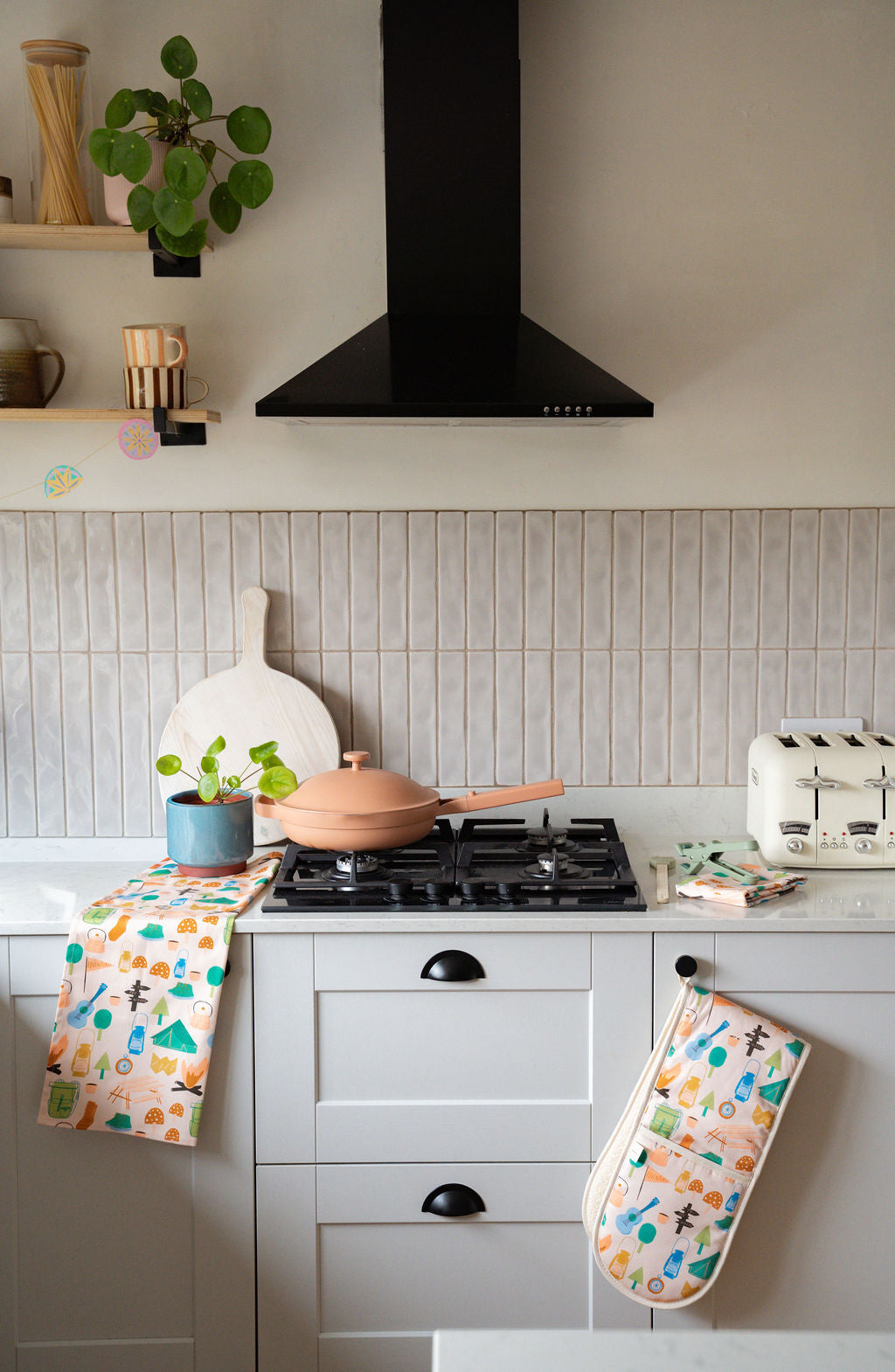 A styled kitchen scene with white cabinets, a black range hood, a beige tiled backsplash, and a terracotta-colored frying pan on the stovetop. A peach oven mitt and a patterned kitchen towel with colorful trees and animals hang from the cabinet handles.