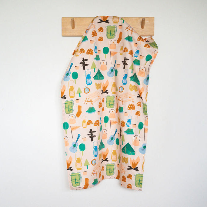 A cotton tea towel colourful camping themed print, hanging from a wooden peg on a plain white wall.