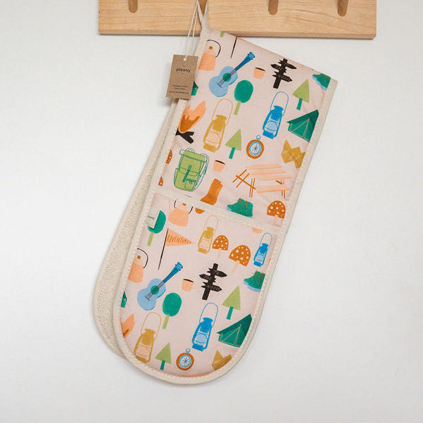 Fabric oven gloves with a colourful pattern of camping items from a wooden wall hook on an off-white wall, with a dark blue grey cabinet visible at the top.
