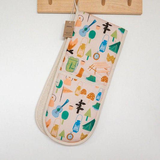 Fabric oven gloves with a colourful pattern of camping items from a wooden wall hook on an off-white wall, with a dark blue grey cabinet visible at the top.
