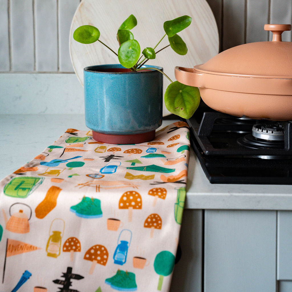 A kitchen countertop scene with a small potted green plant in a teal pot, a large white wooden paddle board leaning behind it, a peach-colored Dutch oven on a black stovetop to the right, and a colorful patterned cloth draped across the counter against a gray tiled wall.