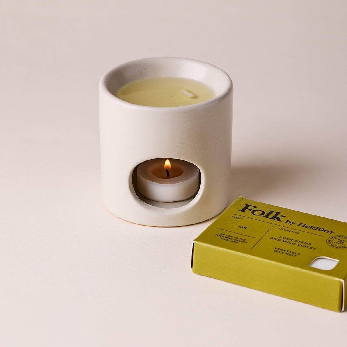 White ceramic candle holder with a lit candle and a yellow box labeled 'Folk' on a beige background