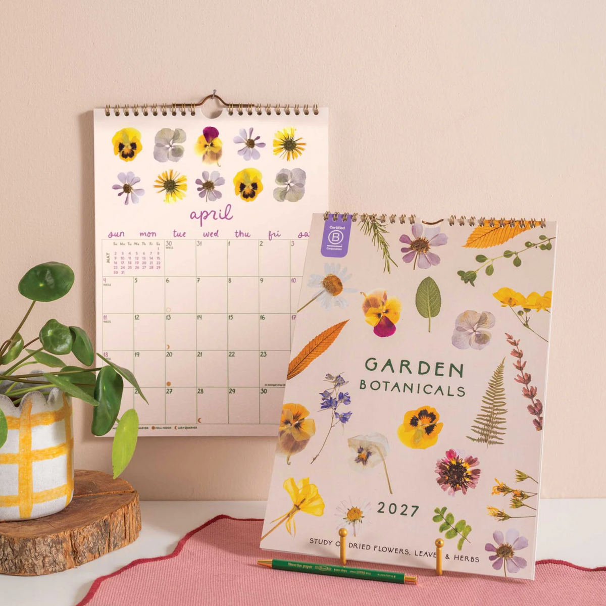 A spiral-bound 2027 Garden Botanicals desk calendar stands on a pink rug next to a matching monthly calendar page, with a small potted plant and wood slice prop on a light beige background.