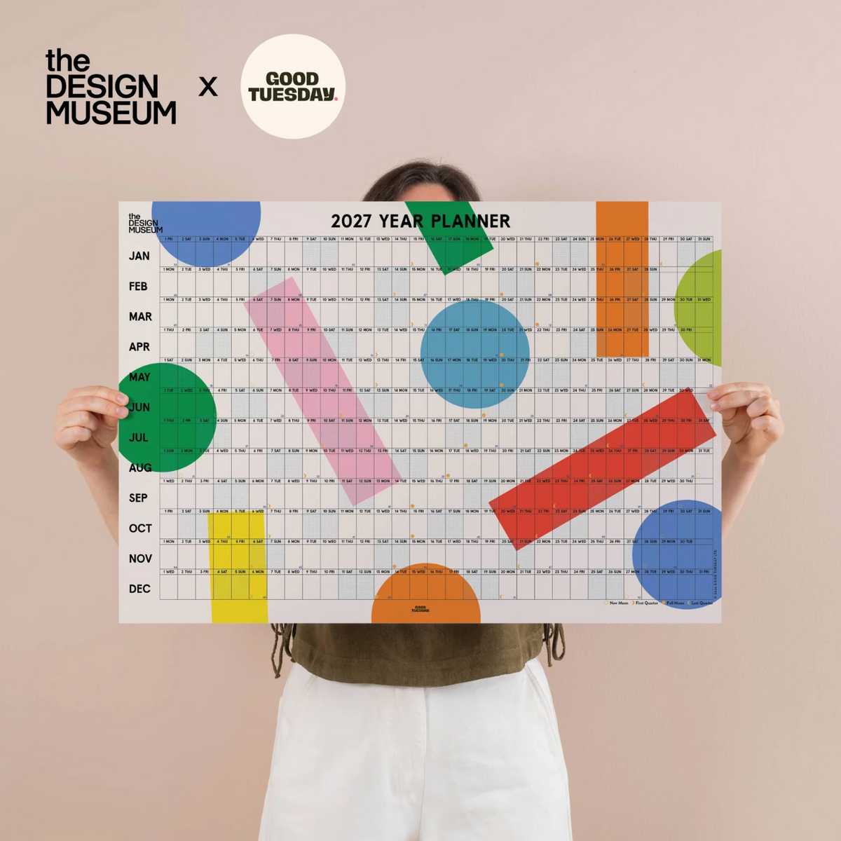 A person holds up a large 2027 year planner poster featuring a grid layout and colorful abstract geometric shapes. The background is a solid pale pink, with 'the DESIGN MUSEUM x GOOD TUESDAY' branding at the top.