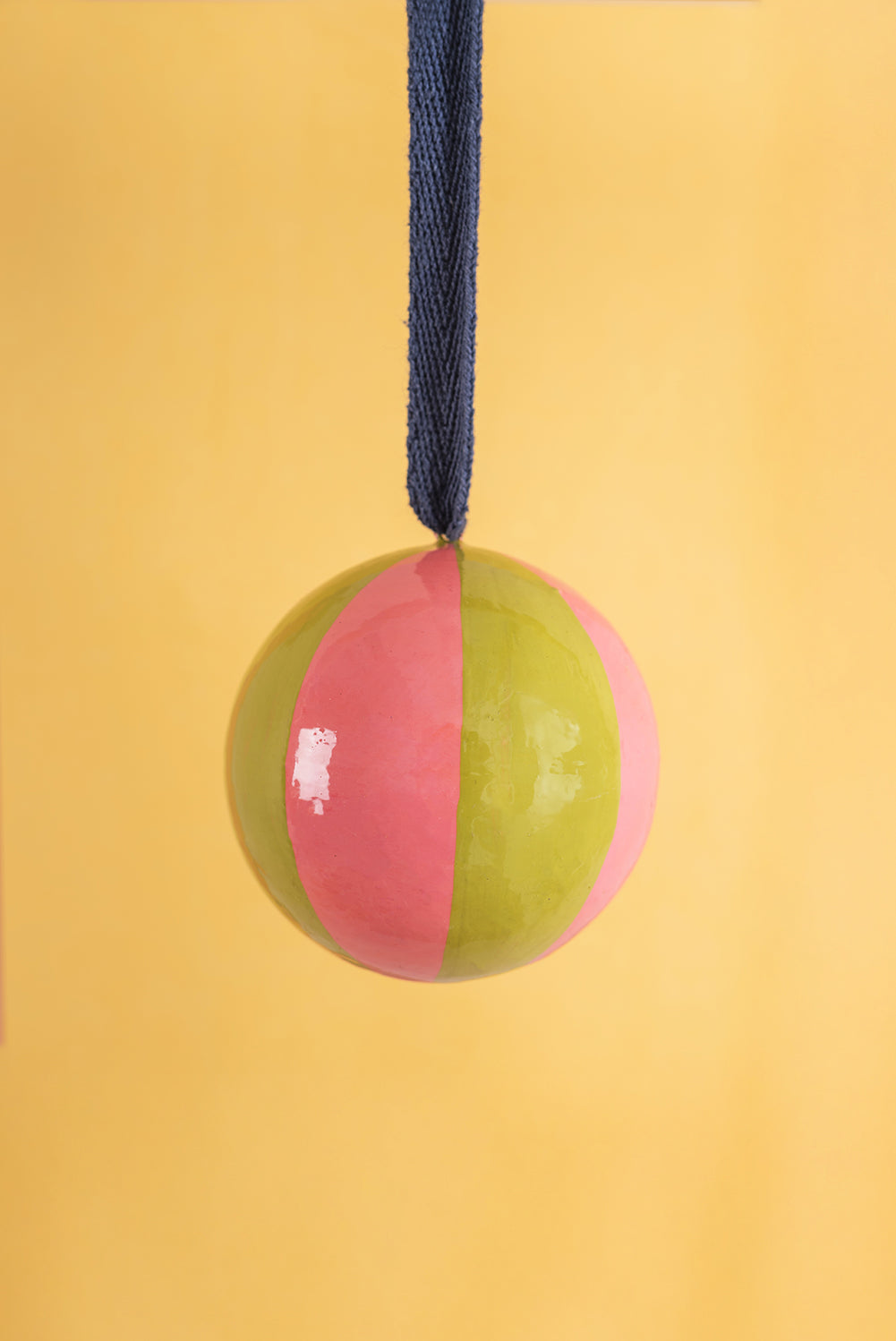 Two-toned pink and green christmas bauble with a blue rope on a yellow background