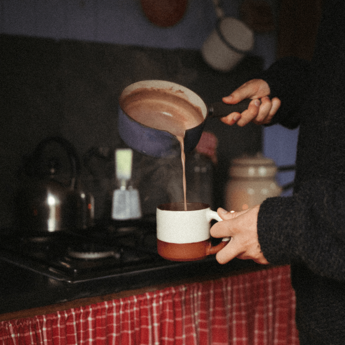Hot chocolate being poured from a saucepan into a mug on a stove.