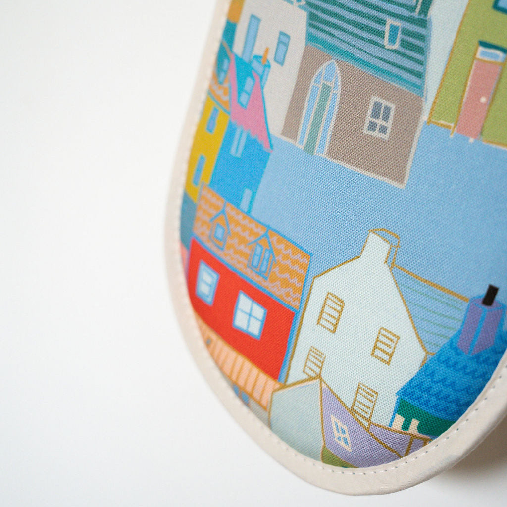 A close-up of cotton double oven gloves, featuring a colourful printed pattern of houses and rooftops in shades of blue, red, yellow, green, and beige.