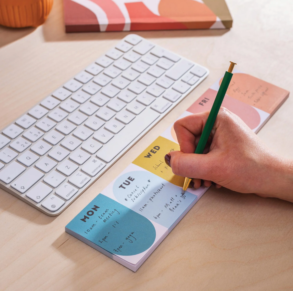 A close-up of a hand writing on a weekly planner sticky note pad with a green and gold pen, placed on a light wood desk next to a white wireless keyboard.