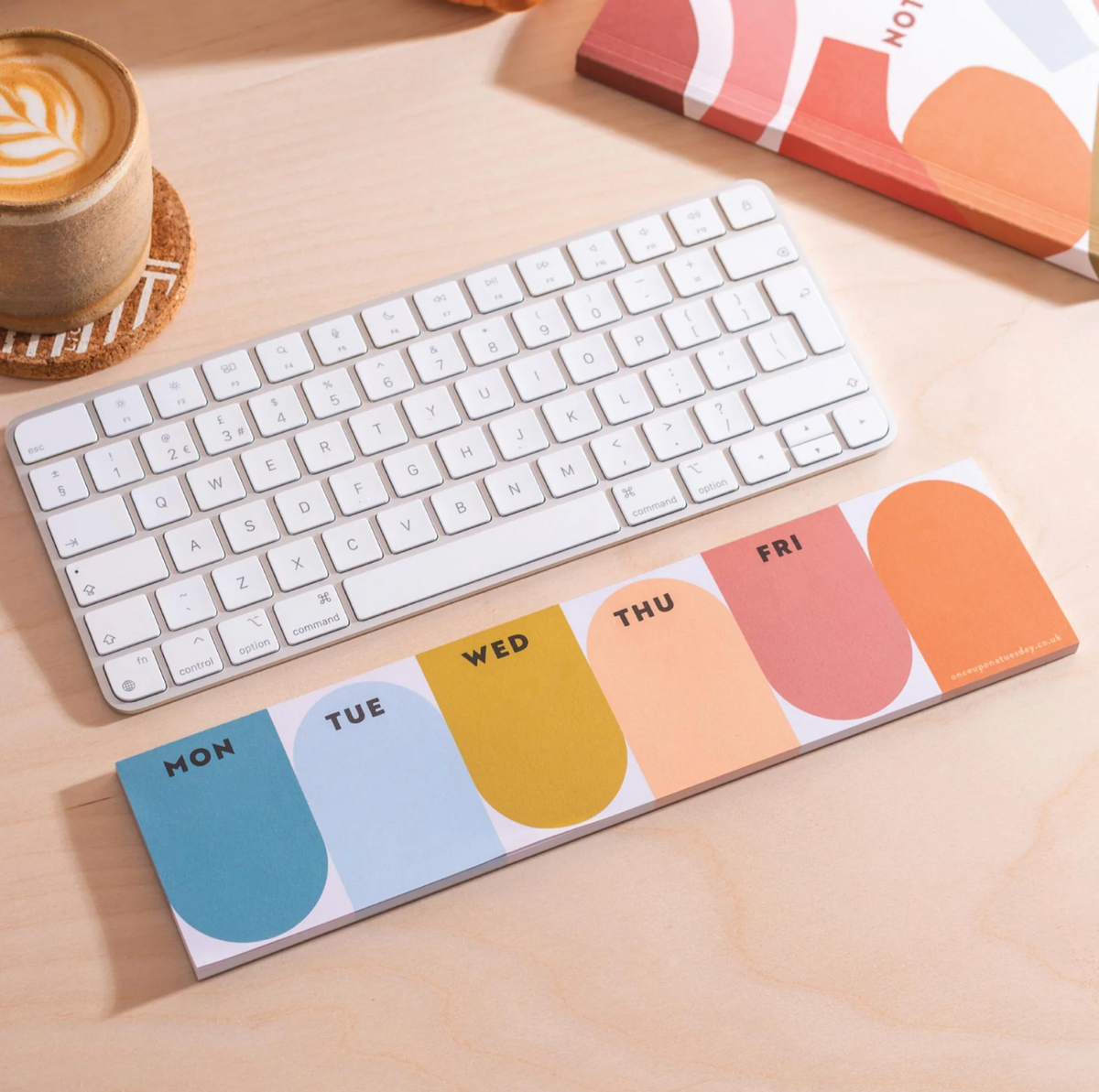 A weekly planner notepad with rounded daily tabs labeled MON through FRI lies on a light wood desk beside a white wireless keyboard. A coffee cup and part of a book are also visible in a styled workspace setting.
