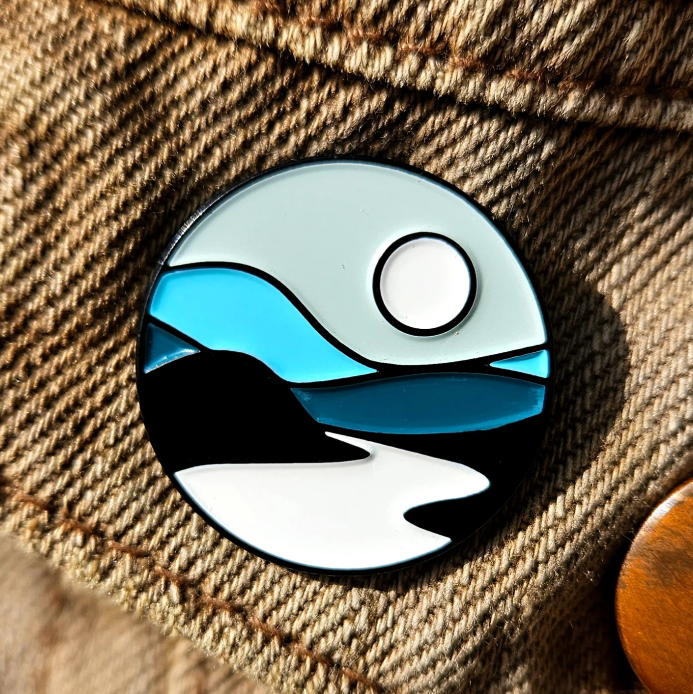 Pin with a design of a landscape featuring water, sky, and mountains on a textured fabric background.