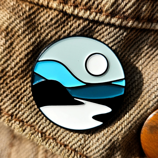 Pin with a design of a landscape featuring water, sky, and mountains on a textured fabric background.