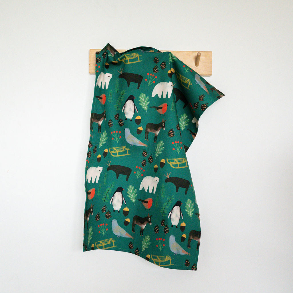 A green fabric tea towel with a whimsical animal print hangs from a small wooden peg on a white wall, with a wall cabinet visible at the top