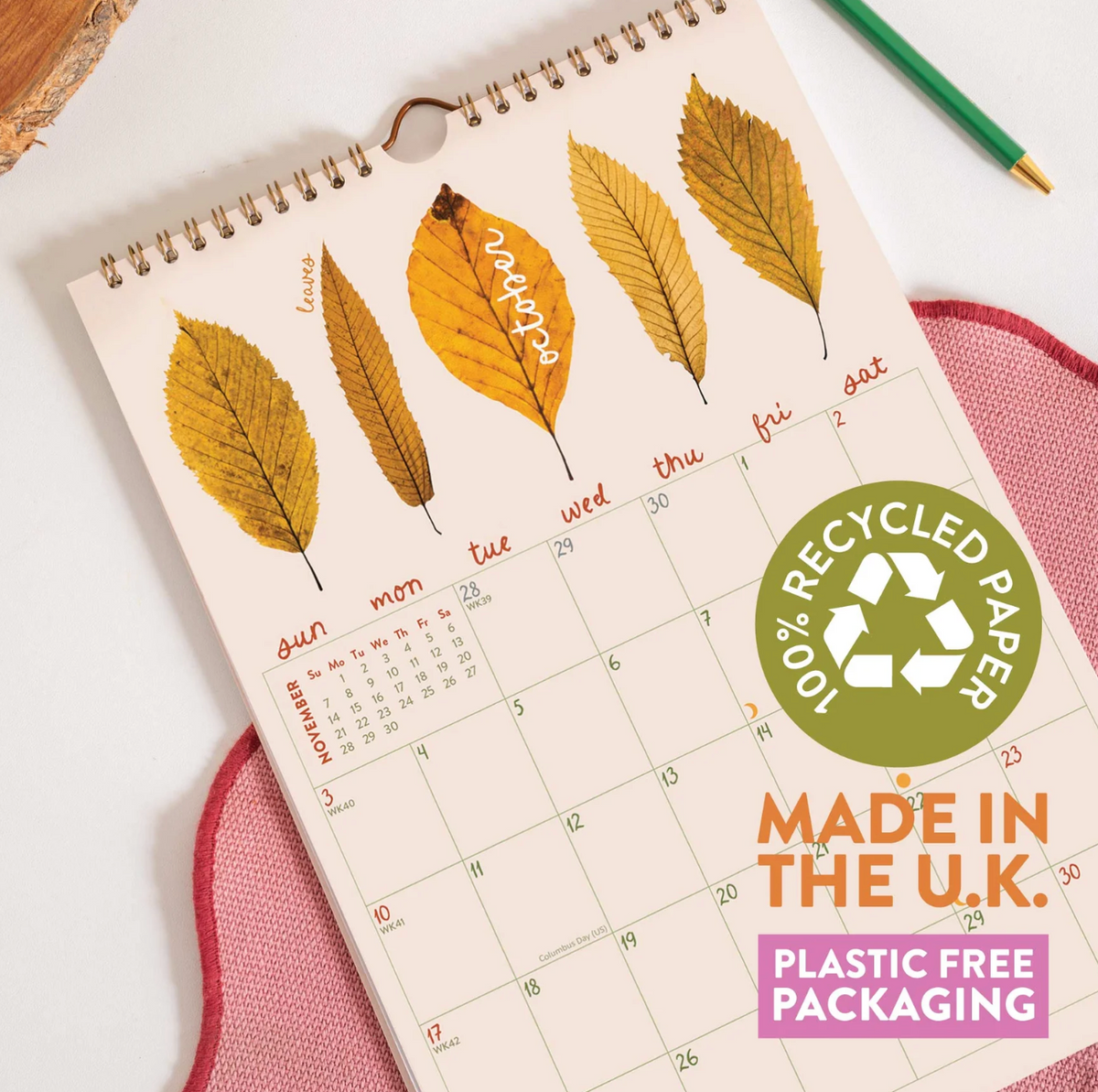 A spiral-bound desk calendar page with autumn leaf illustrations across the top and a monthly calendar grid below. The page is styled on a pink fabric surface with a green pencil nearby, and promotional text on the right reads '100% Recycled Paper' and 'Made in the U.