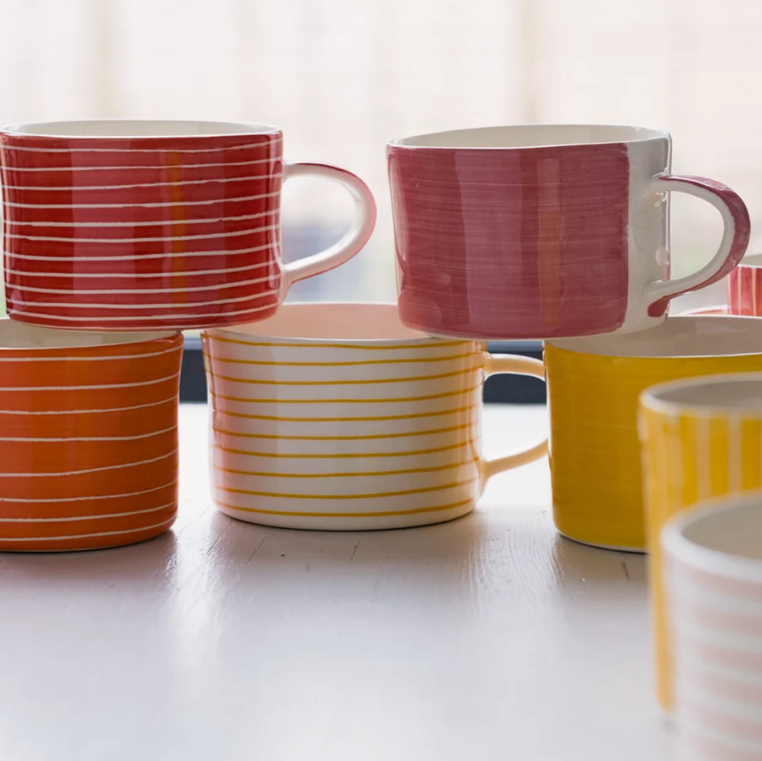 A group of ceramic mugs in assorted colors and stripe patterns, stacked together on a light tabletop with a soft, bright background.