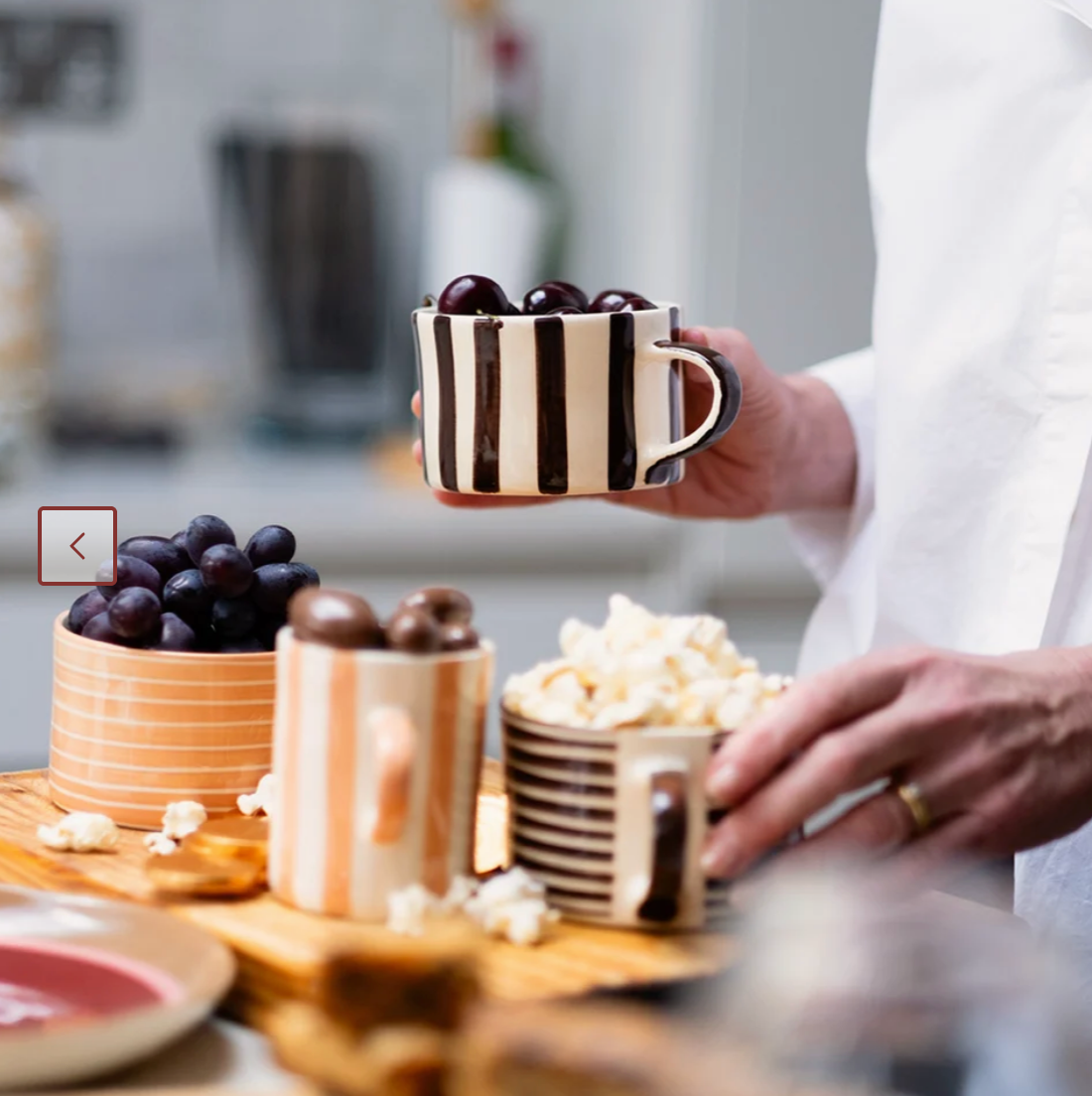 A person in a white shirt holds a striped ceramic mug filled with dark grapes in a kitchen setting, with other patterned mugs and food items on a wooden board in the foreground.