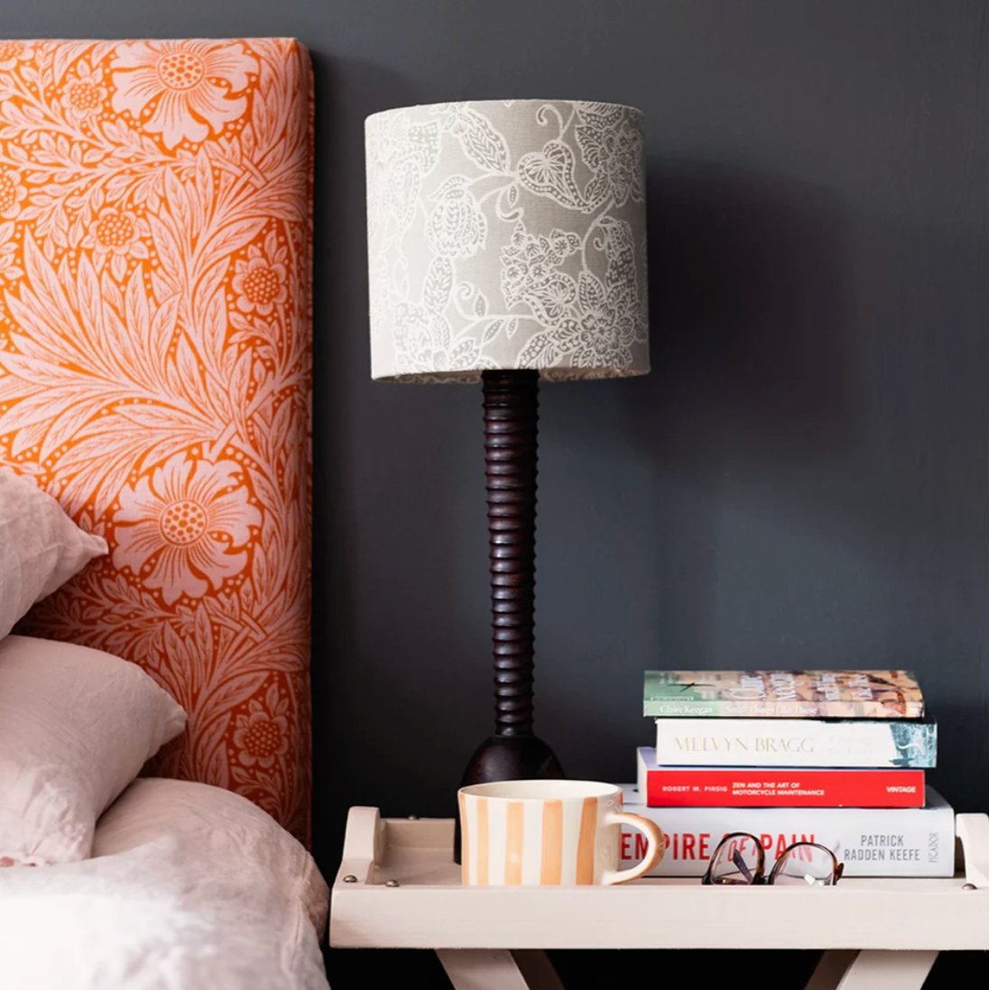 A table lamp with a gray and white damask-patterned drum shade and a dark ribbed stem, styled on a white side table next to a bed with an orange floral headboard against a dark gray wall.