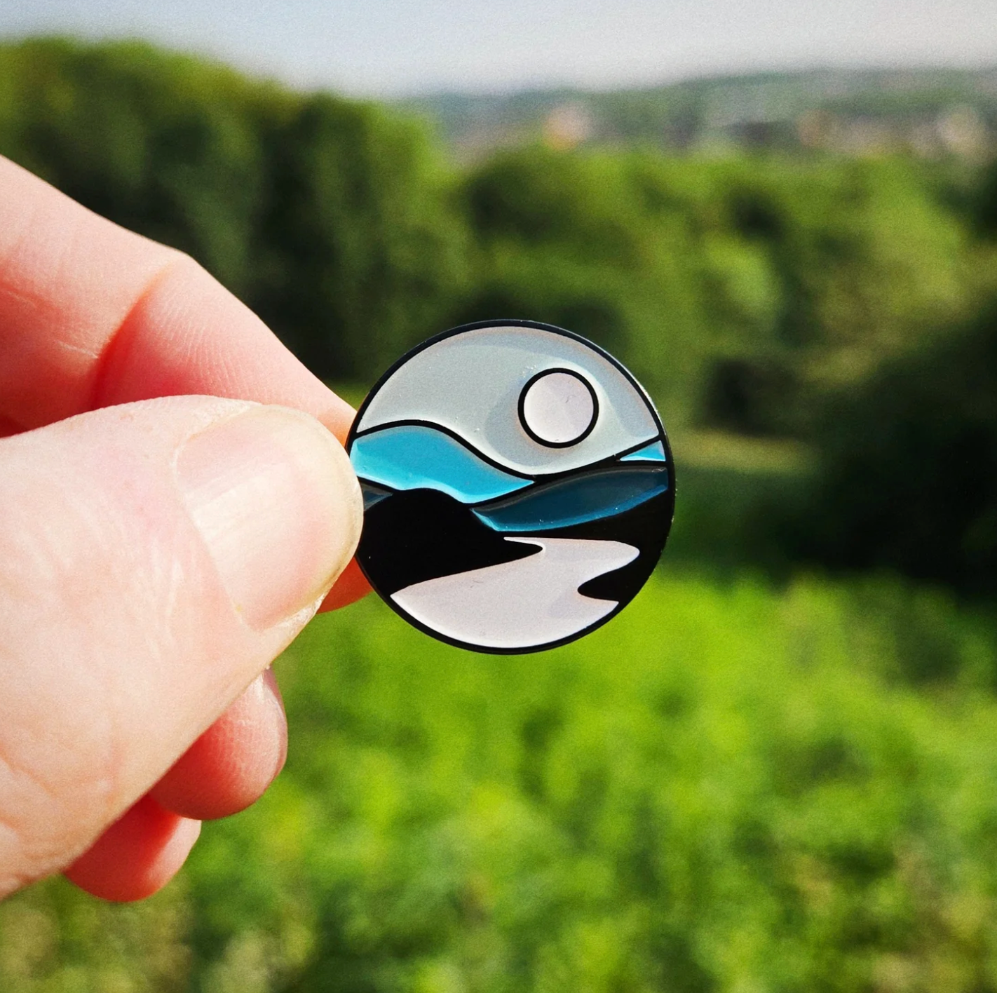 Hand holding a circular badge with a nature design against a blurred green landscape