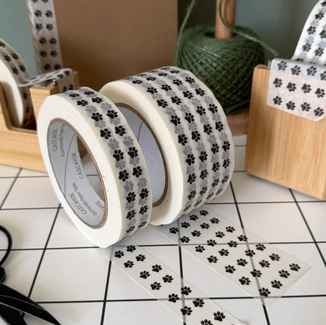 Three rolls of washi tape with a black paw print pattern on a white background, displayed on a white tiled surface with other rolls and a spool of twine in the background.