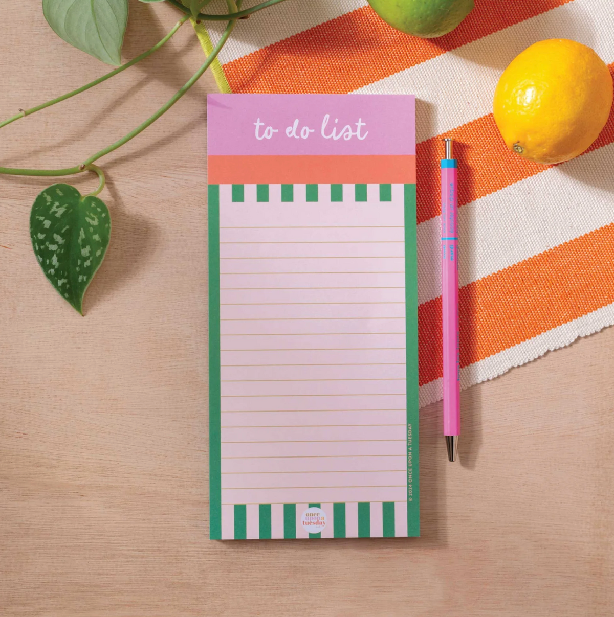A pink and green to-do list notepad with a matching pink pen, styled in a flat lay on a light wood surface with a striped orange and white cloth, green leaves, and a lemon.