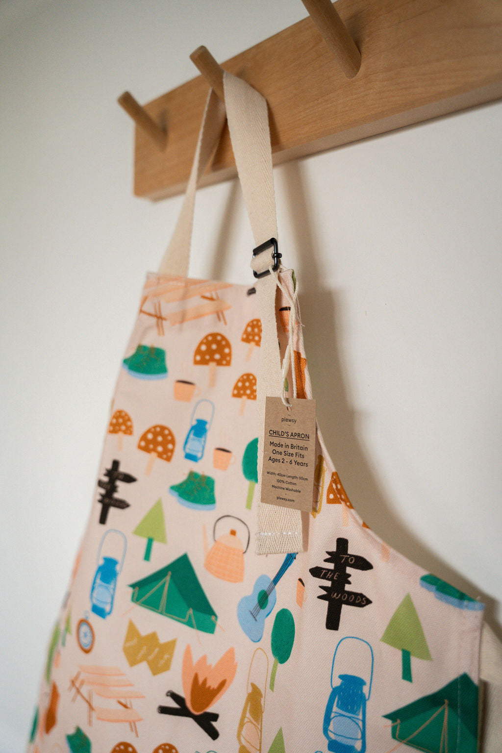 A cream-coloured canvas apron with a colorful camping-themed print, hanging from a wooden peg on a white wall.