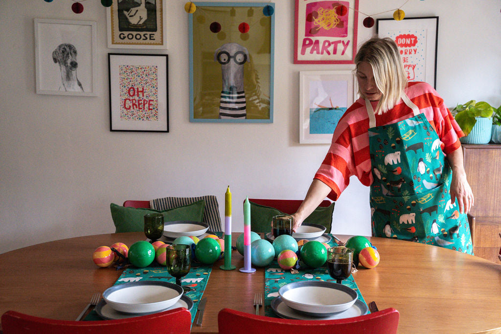 A woman wearing a red and white striped shirt and a green Christmas themed apron with a polar bear pattern is setting a table with colourful christmas baubles, placemats, and candles in a decorated room with framed art.