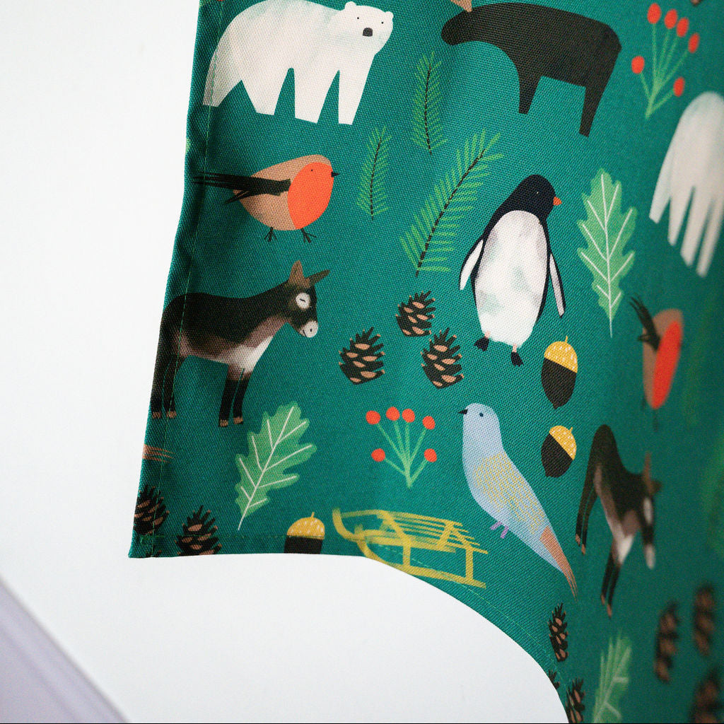 A close up of green apron fabric featuring a whimsical winter animal print with polar bears, penguins, birds, pinecones, acorns, and evergreen leaves, shown against a white background.