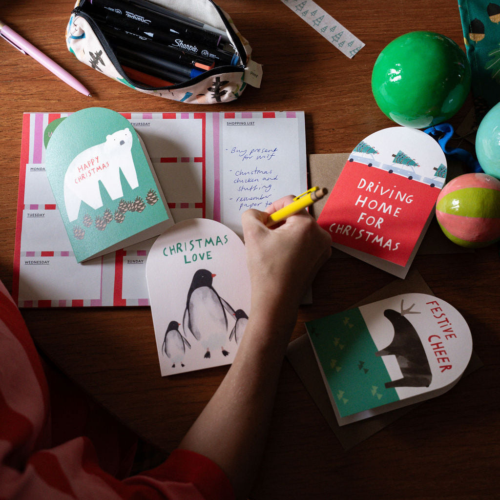 A person's hand is writing in a notebook on a wooden desk, surrounded by Christmas-themed stationery, including greeting cards with polar bear and penguin illustrations, a patterned pencil pouch, washi tape, and colorful ornaments.