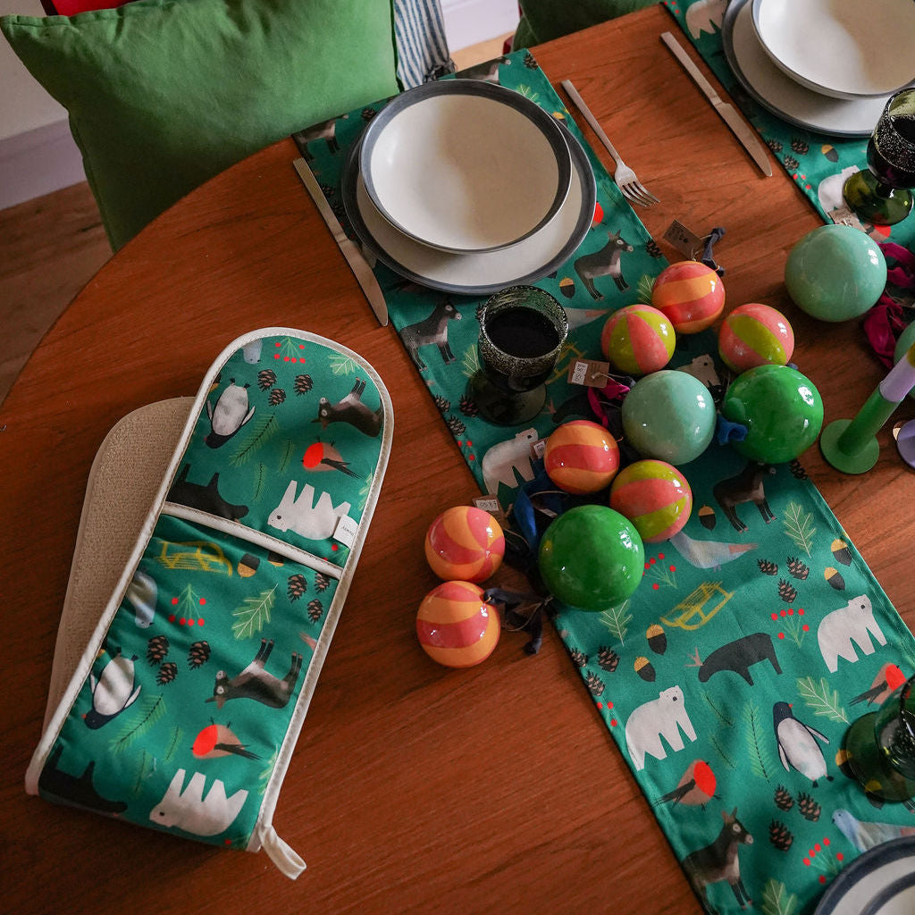 A green table runner and matching double oven gloves with a whimsical polar bear print, styled on a wooden dining table with plates, colorful ornaments, and green pillows on red chairs.