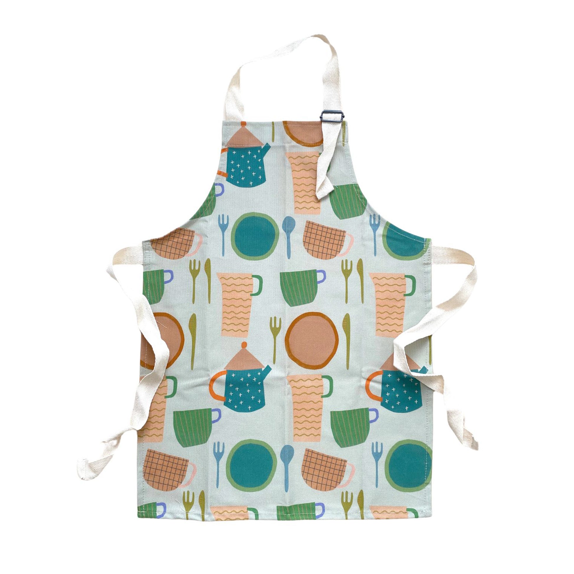 A children's apron with a tea time theme, featuring illustrations of cups, plates, and cutlery in a multicolor design on a white background.