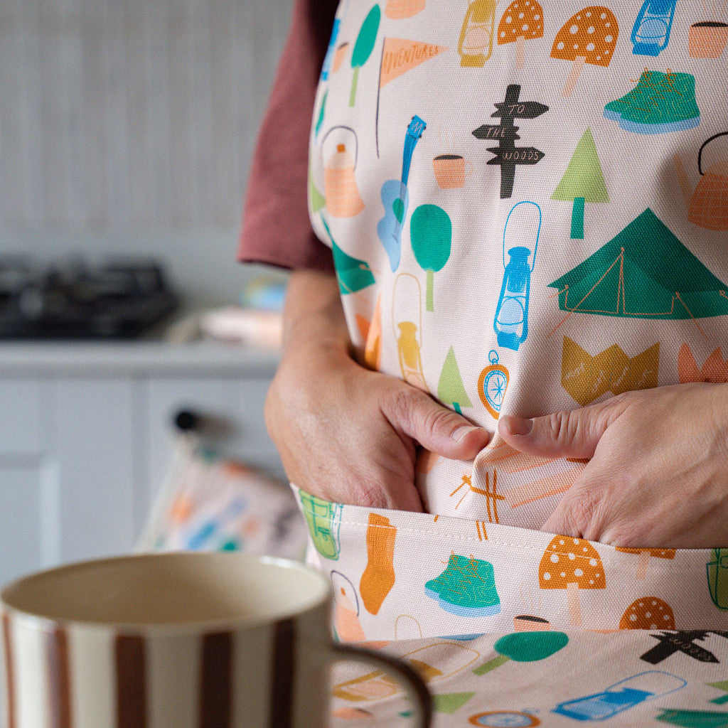 A person wearing a cream-coloured apron with a colorful camping-themed print, including tents, trees, and lanterns. The person is shown from the chest down, with their hands adjusting the waist apron.