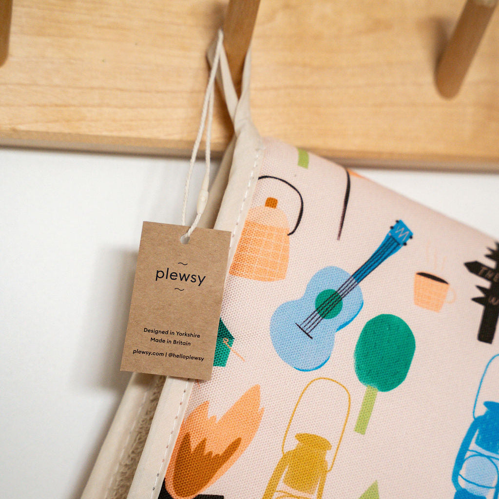 A close-up of a plewsy explore double oven gloves with a light pink background and a colorful outdoor-themed print including a blue guitar, green trees, yellow lantern, and orange campfire. A kraft paper tag with the 'plewsy' brand is hanging from the fabric.