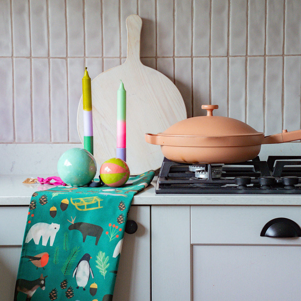 A colourful kitchen scene featuring a green kitchen towel with a whimsical animal print hanging from a white drawer, next to a terracotta-coloured pan on a gas stovetop. A wooden paddle board leans against a beige tiled backsplash in the background.