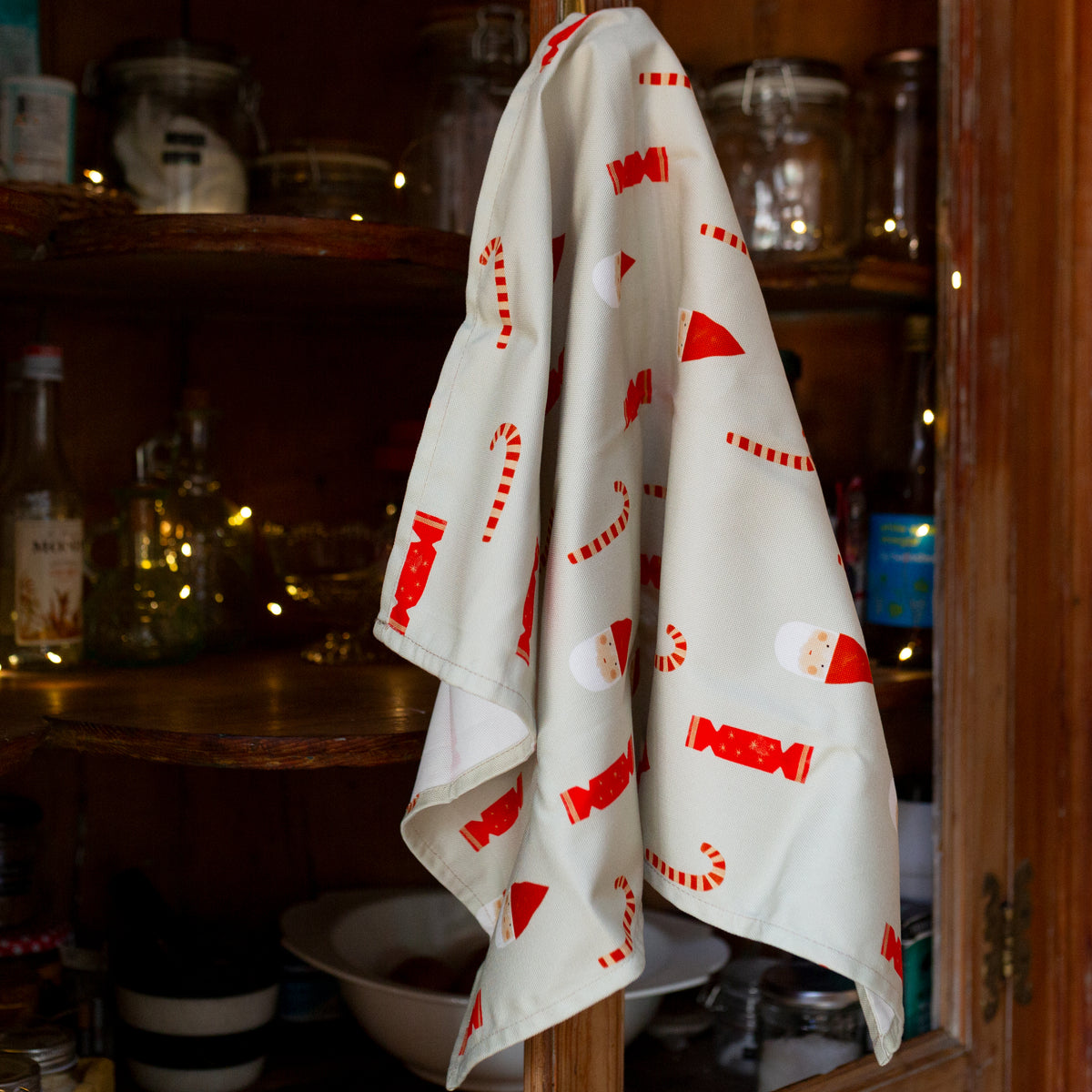 An off-white cotton tea towel featuring a festive Christmas pattern of red and white candy canes, Santa Claus faces, and red Christmas ornaments.